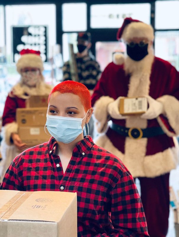 Masked shoppers in a store carry cardboard boxes, with two Santa figures in the background.