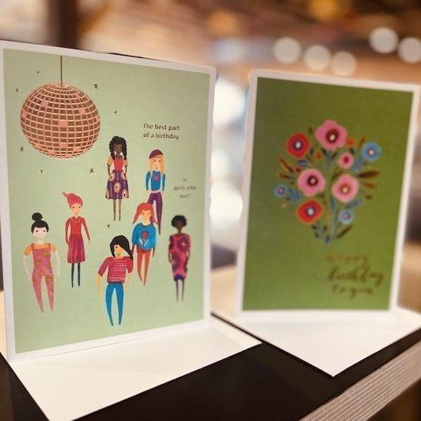 Two standing greeting cards on a table, one with illustrated people under a lantern, the other with a floral bouquet.