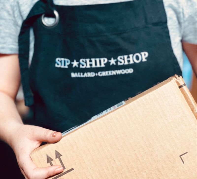 Person holding a cardboard box in front of a black apron with “Sip Shop Shop” text