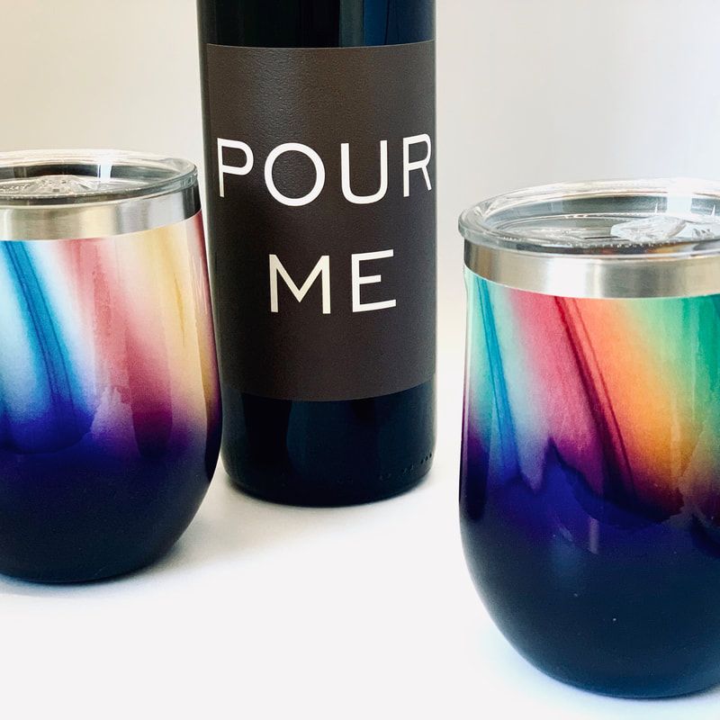 Two iridescent stemless wine glasses beside a black bottle labeled “POUR ME”