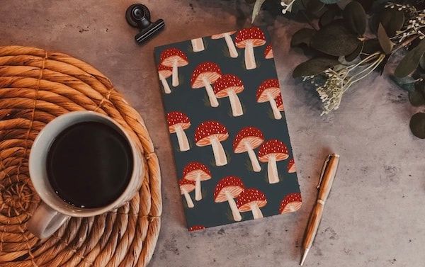 Coffee, mushroom-pattern notebook, pen, and dried flowers on a rustic desk surface.