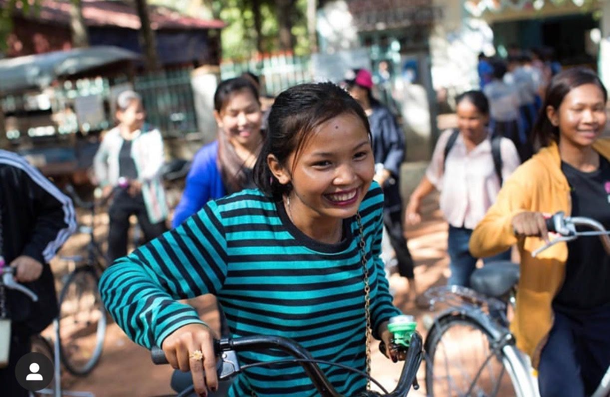 Woman in a turquoise striped shirt smiling and riding a bicycle in a busy outdoor market.