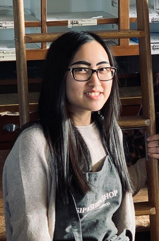 Woman with long black hair and glasses, smiling indoors against a wooden staircase.