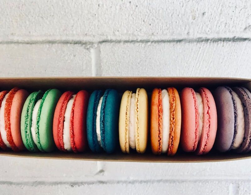 Colorful macarons arranged in a row inside a narrow box against a white brick background
