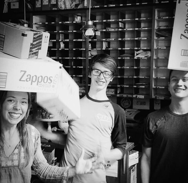 Three smiling workers carrying Zappos boxes in a warehouse shipping area