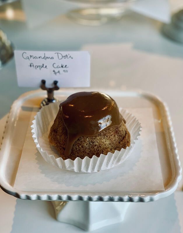 Small caramel-topped dessert cup labeled “Cinnamon Dolce Apple Cake” on a white platter stand.