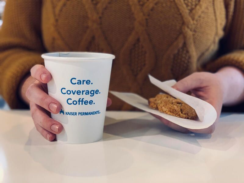 Hands holding a white cup and a cookie on a table, with “Care. Coverage. Coffee.” printed on the cup
