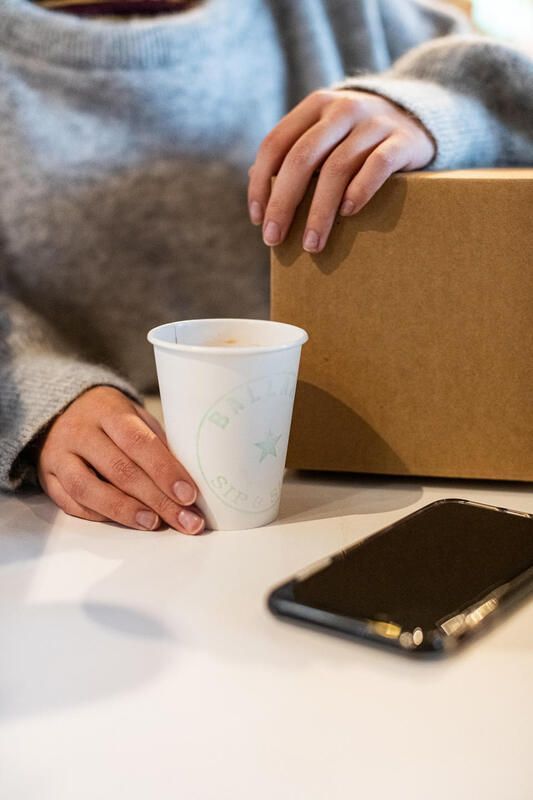 Hands holding a paper cup and cardboard box beside a smartphone on a white table.