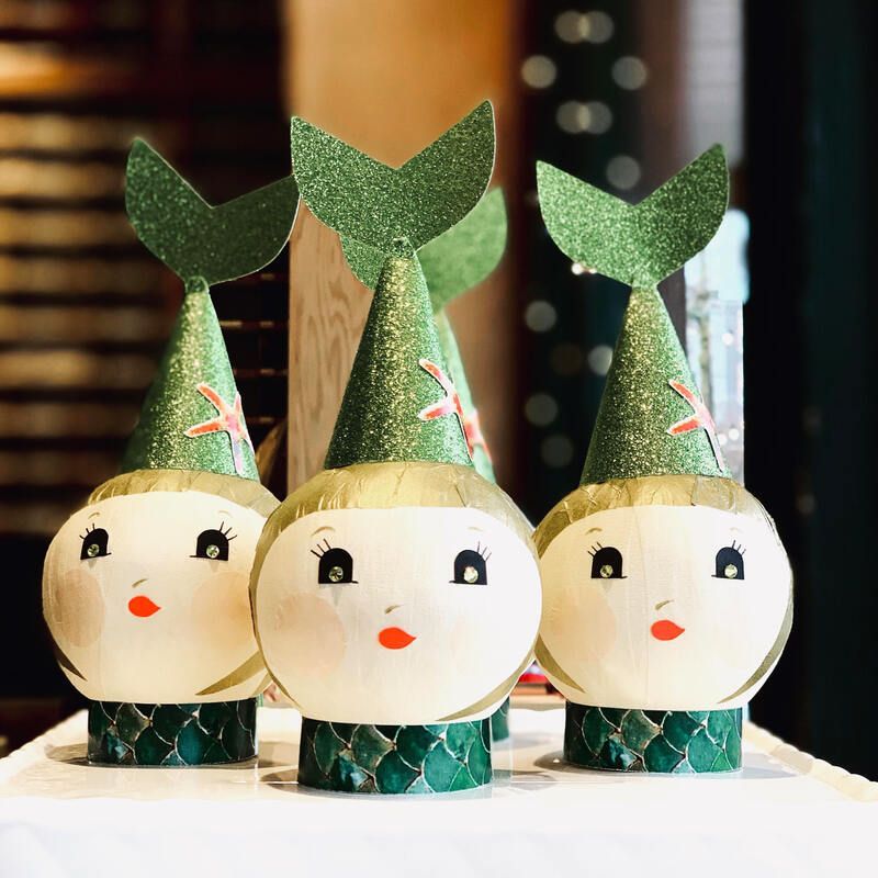 Three green fish-shaped holiday crafts with round faces and paper hats on a table
