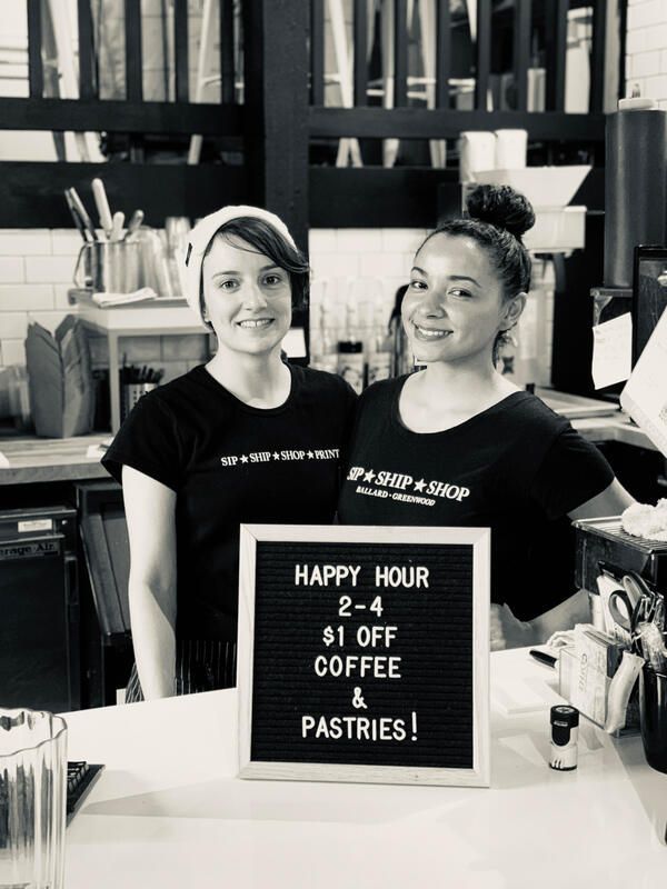 Two café staff stand behind a sign advertising happy hour coffee and pastries.