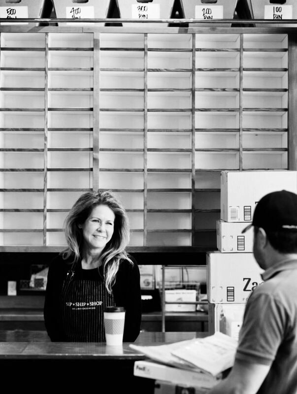Woman behind café counter smiling at a customer, with stacked boxes and shelves in the background.