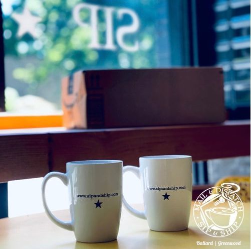 Two white mugs on a wooden table by a window, with a blurred café sign in the background.