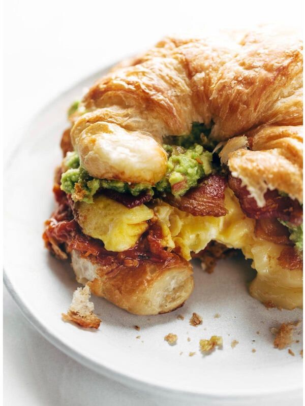 Croissant sandwich with egg, bacon, and avocado on a white plate, one bite missing