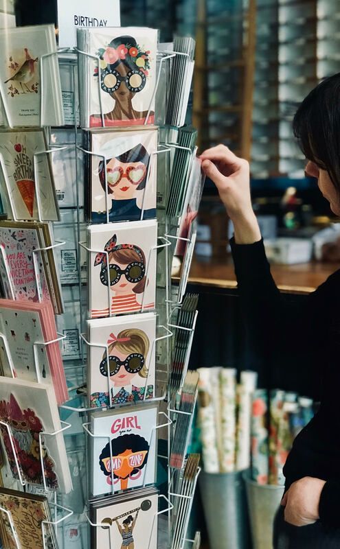 Greeting card rack with colorful illustrated cards; a person reaches toward one card in a shop.