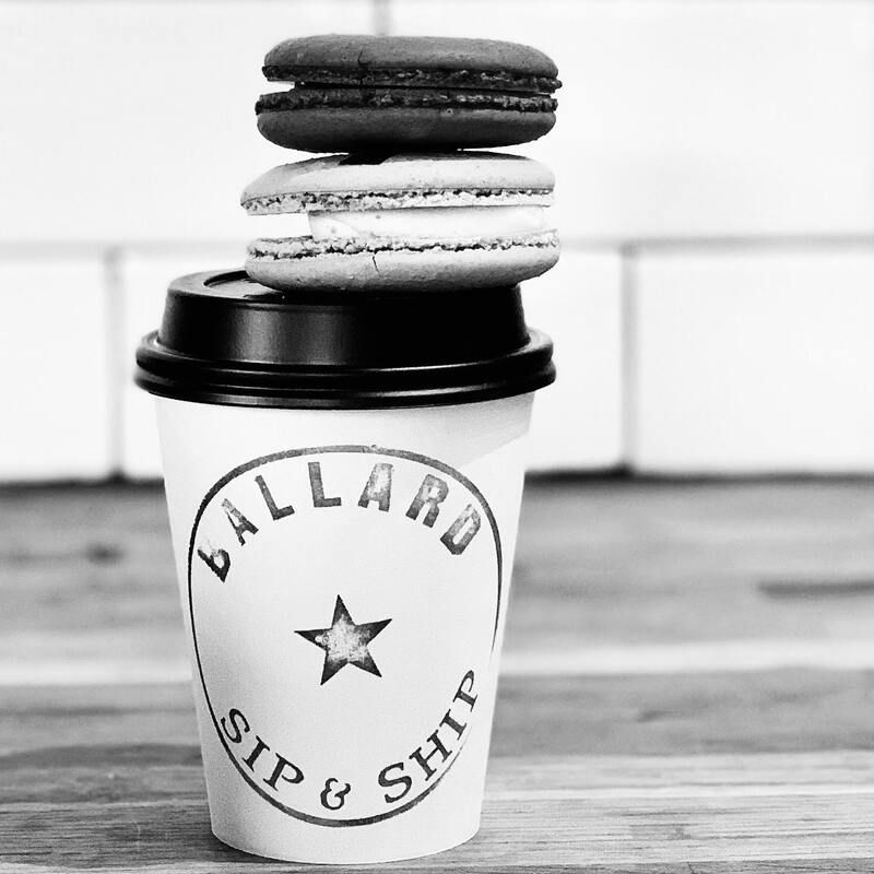 Black-and-white cup with stacked macarons on lid, labeled “Ballard Sip & Ship” in a cafe setting