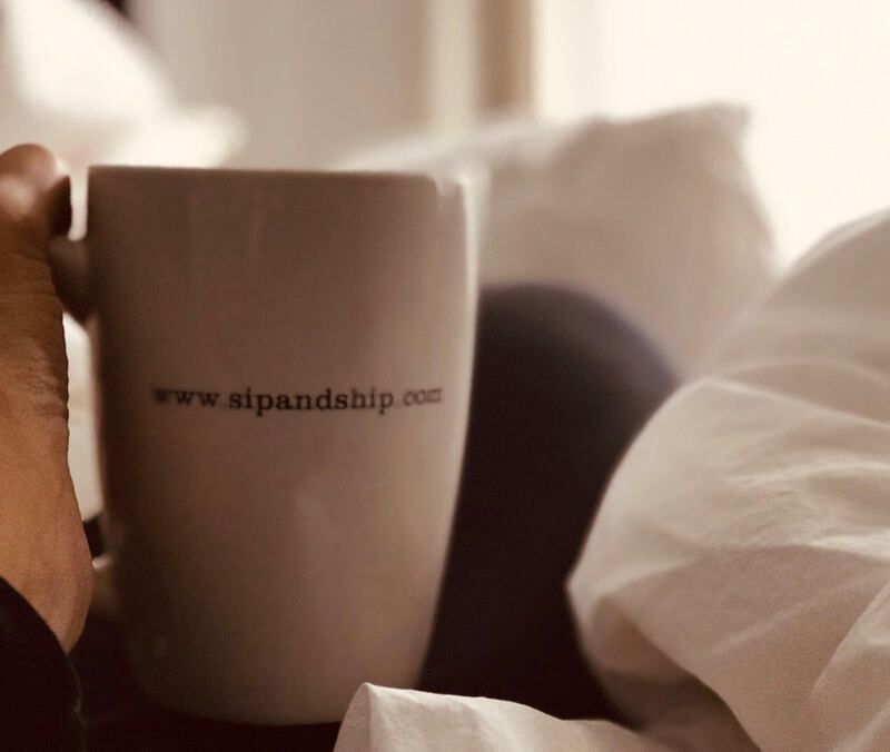 White mug with website text on a bed, beside a hand and soft pillows in warm light