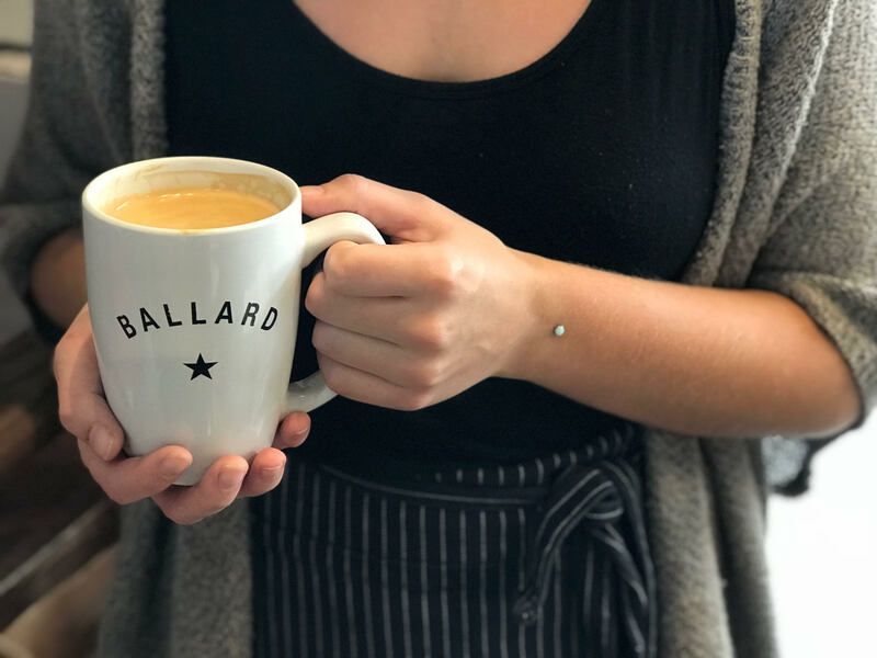Person holding a white Ballard mug with a drink, wearing a gray cardigan and black top.