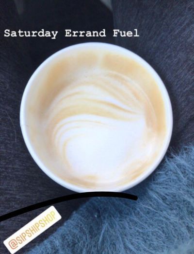 Latte in a white cup with foam art on a dark table, with text reading “Saturday Errand Fuel.”