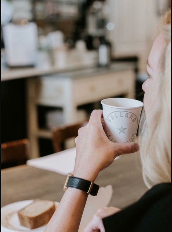 Person holding a white cup in a café, with a slice of bread on the table.