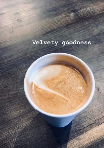 Latte in a white cup on a wooden table, with “Velvety goodness” text above.