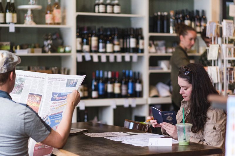 Customers seated at a cafe counter, one reading a newspaper and another using a phone, with shelves of bottles behind them