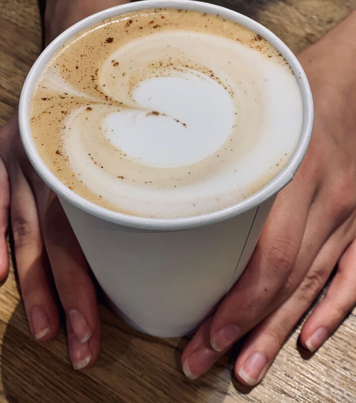 Hands holding a white paper cup of latte with foam art on a wooden table.