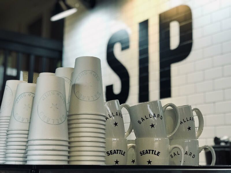 Stacks of white Seattle mugs and paper cups on a counter in front of a SIP sign.
