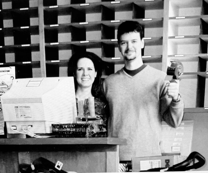 Two people behind a counter, one holding a can, in front of shelves and a cash register.