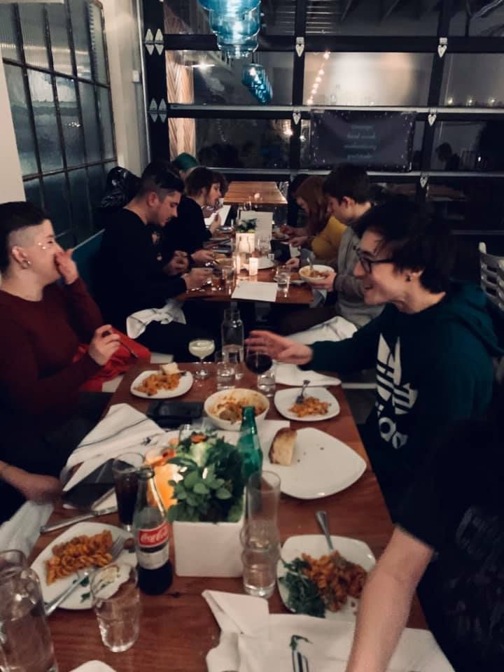 Friends dining at a long table in a dimly lit restaurant, sharing plates and drinks together.