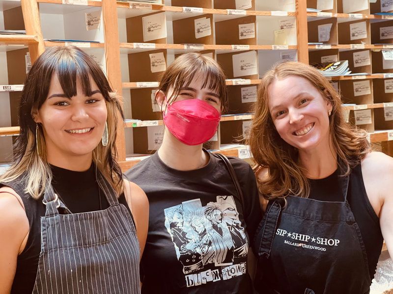 Three people smiling indoors in front of wooden shelves, one wearing a pink face mask