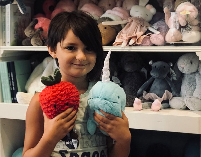 Child holding two plush fruit toys in front of a shelf of stuffed animals