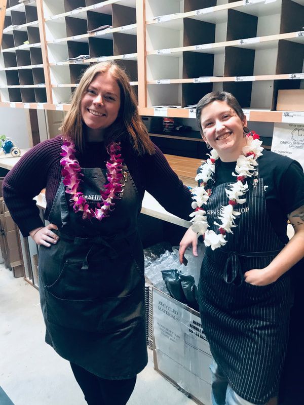 Two people smiling in aprons and floral leis, posing in front of cubby shelves in a workspace
