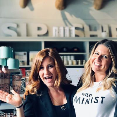 Two women smiling and waving in a boutique with shelves of products and a “SPRING” sign behind them