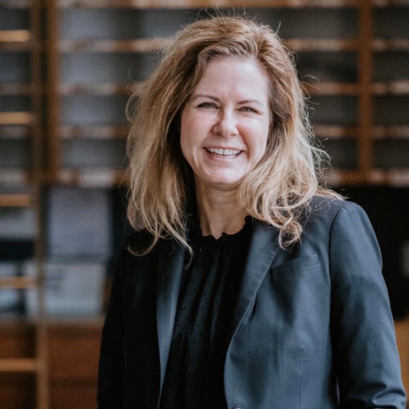 Smiling woman in a black blazer standing indoors with shelves blurred in the background