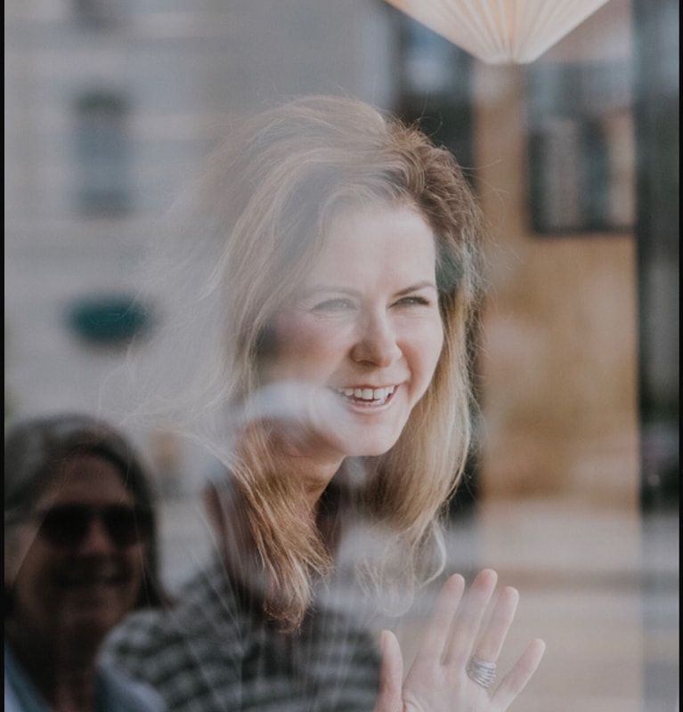 Smiling woman seen through a window, with a blurred reflection in the foreground.