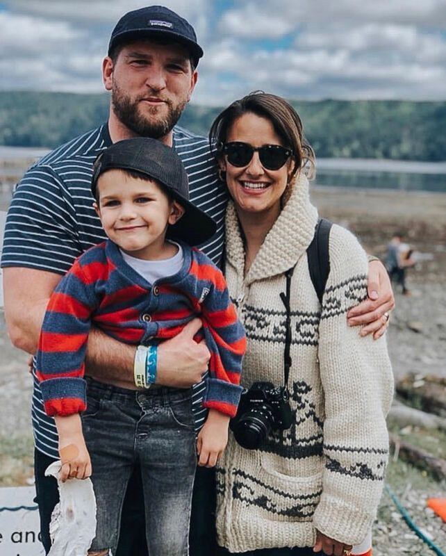 Three people posing outdoors by a waterfront, with mountains and cloudy sky in the background