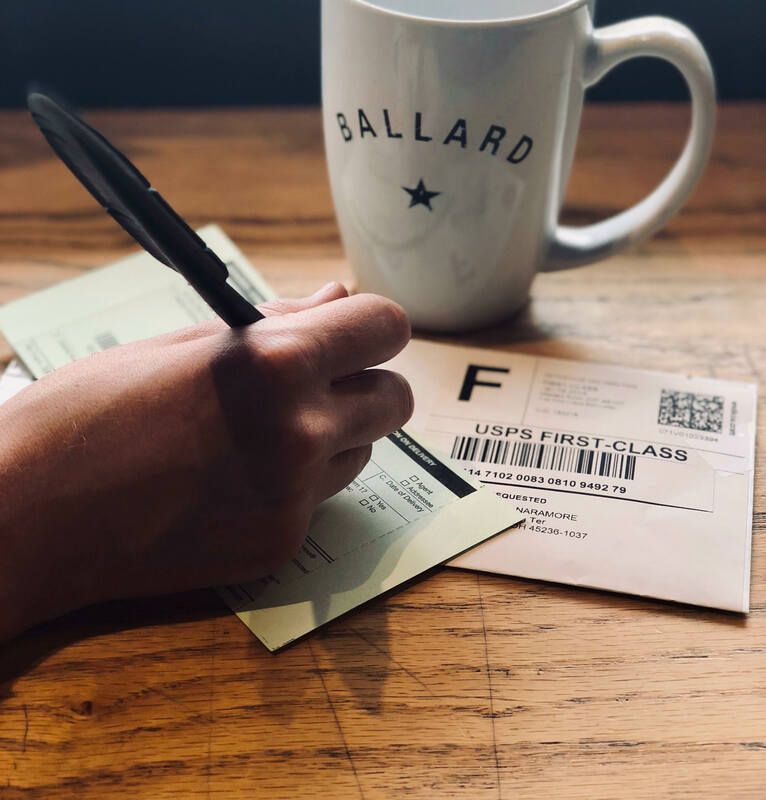 Handwriting a USPS shipping label beside a white Ballard mug on a wooden table