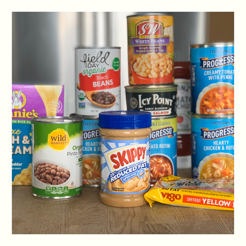 Assorted canned and jarred foods on a wooden table, including beans, tuna, soup, and peanut butter.