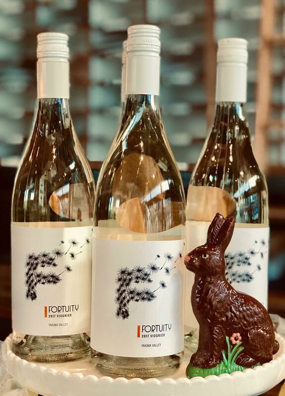 Three white wine bottles with labels, beside a chocolate bunny and small flowers on a rustic surface.