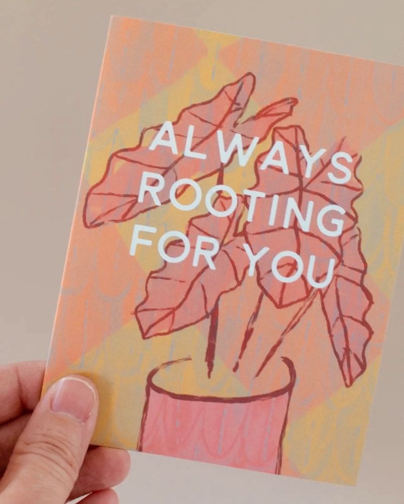 Hand holding a card with pink leaves in a pot and the words “ALWAYS ROOTING FOR YOU”