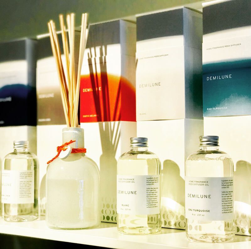 Fragrance bottles and reed diffusers on a sunlit shelf with white, red, and blue packaging