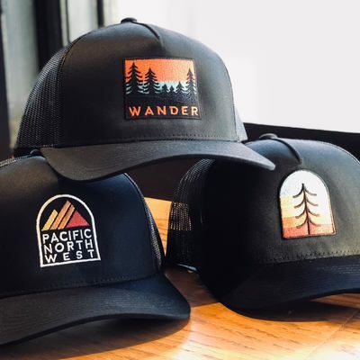 Three black trucker hats with outdoor-themed patches on a wooden surface