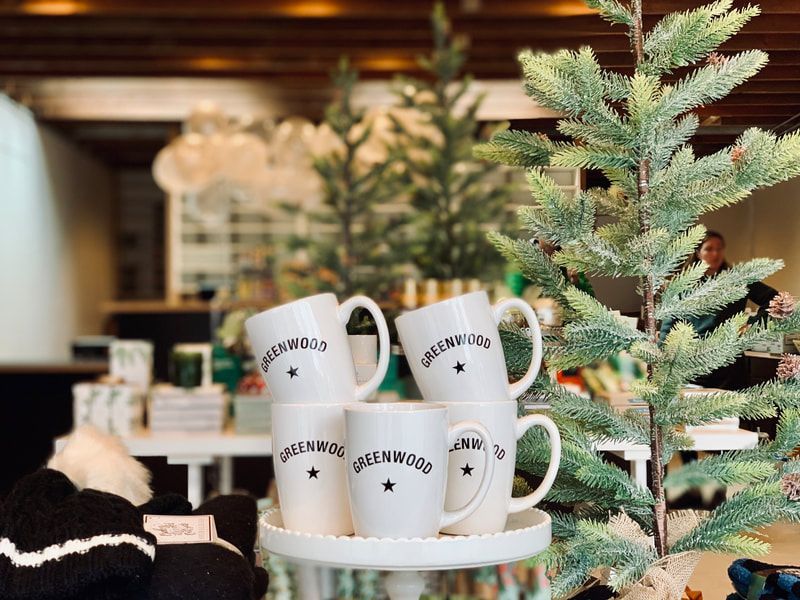 Stacked white mugs with black text beside a small frosted evergreen in a cozy shop display
