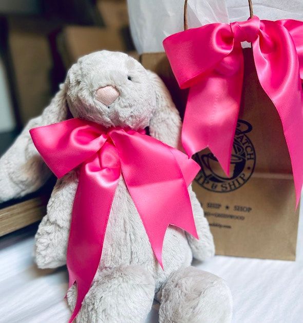 White plush bunny with bright pink bows beside a gift bag on a bed.