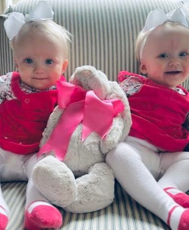 Two white-clad dolls in pink outfits sit on a striped couch beside a stuffed bunny with a pink bow.
