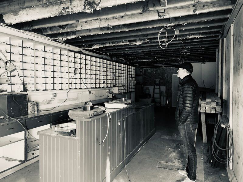 Person standing in a dim industrial room with exposed beams, a long counter, and shelves of supplies.