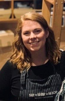 Smiling person in a black shirt and striped apron standing indoors among wooden shelves.