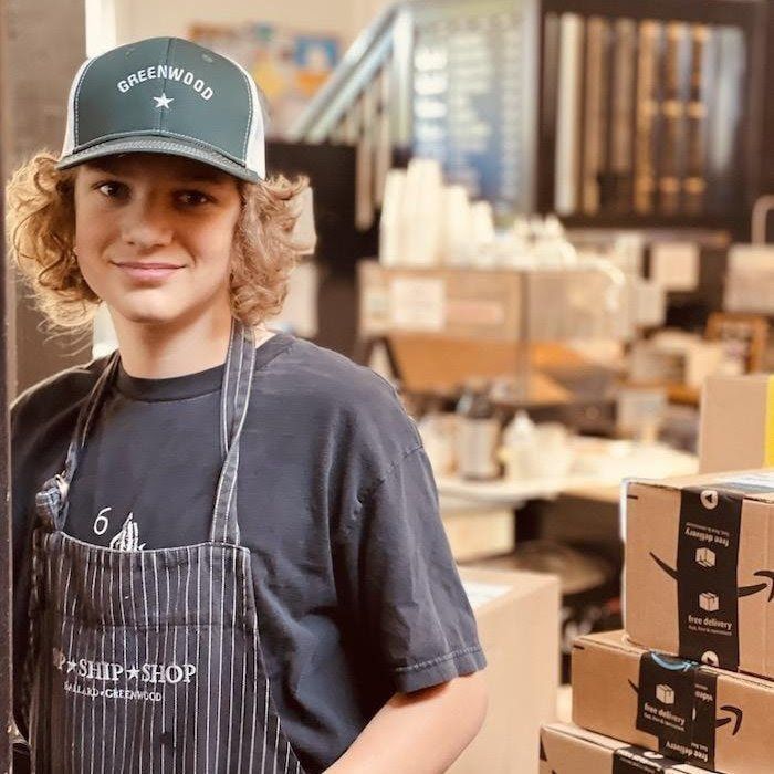 Person in apron and cap stands in a coffee roastery among stacked boxes.