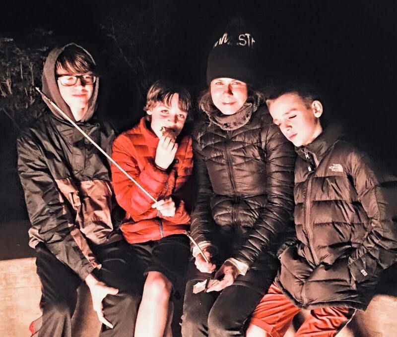 Four friends posing together at night by a campfire, wearing winter jackets and beanies.