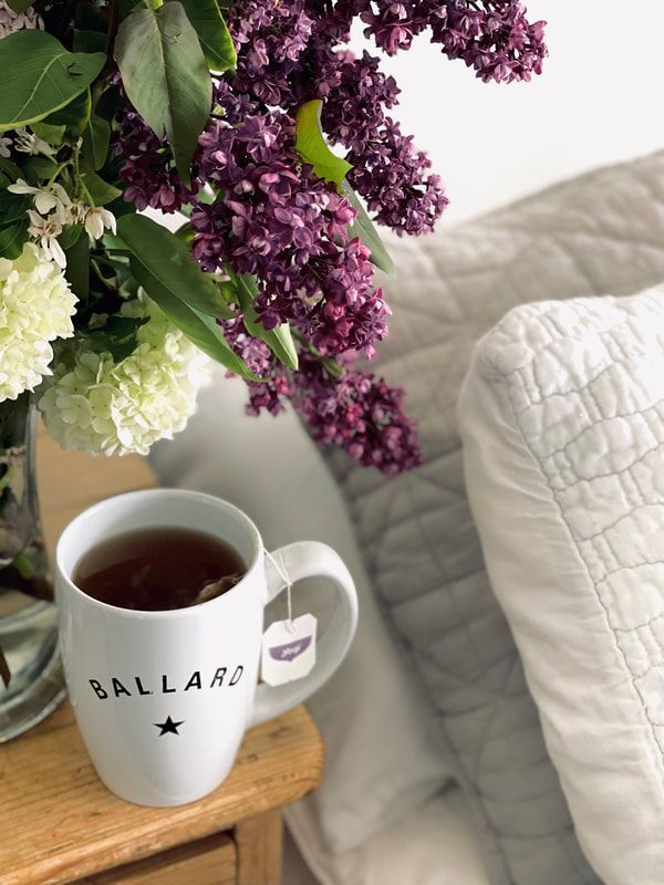 Coffee mug on a wooden table beside a vase of purple and white flowers, near a white quilted chair